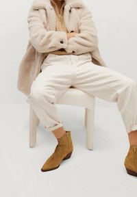 Personne assise, les jambes écartées, portant une veste en fausse fourrure beige, un pull à fermeture éclair beige clair, un pantalon blanc cassé et des bottines en daim beige à talons bas.