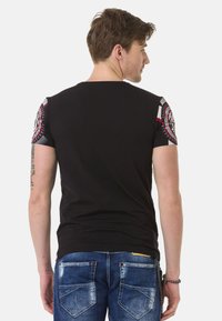 Schwarzes T-Shirt mit kurzen Ärmeln und gemusterten grauen und roten Akzenten an den Schultern. Weiches Material, figurbetontes Design und keine sichtbaren Logos oder Texte.