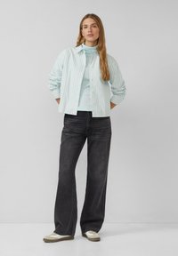 Vrouw staat met haar handen achter haar rug, gekleed in een lichtblauwe gestreepte blouse, een bijpassende coltrui, zwarte wijde jeans en witte sneakers.