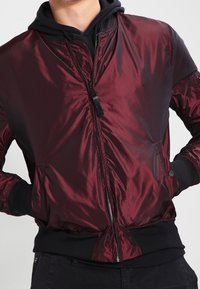 Bordowa kurtka bomberka wykonana z błyszczącego materiału, zapinana na suwak z przodu, z ściągaczami na mankietach i dole, ozdobiona czarnymi akcentami oraz dwoma bocznymi kieszeniami.