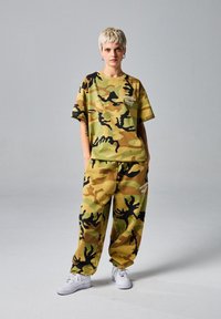 Camiseta y pantalones de corte holgado con estampado de camuflaje en verdes y marrones, con detalles en negro y un logo en el pecho. Combinado con zapatillas blancas.