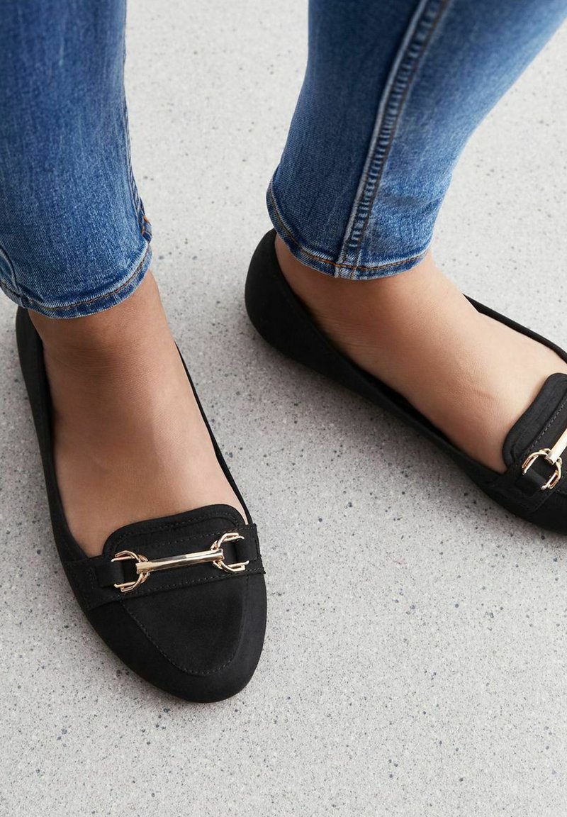 Zwarte ballerina's met een puntige neus, van suede materiaal, met een gouden hardware-accent en een platte zool, gedragen met blauwe spijkerbroeken.