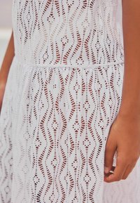 Robe blanche en dentelle avec un motif ondulé et des accents circulaires, présentant une taille froncée et un tissu texturé. Détails visibles sur les manches.
