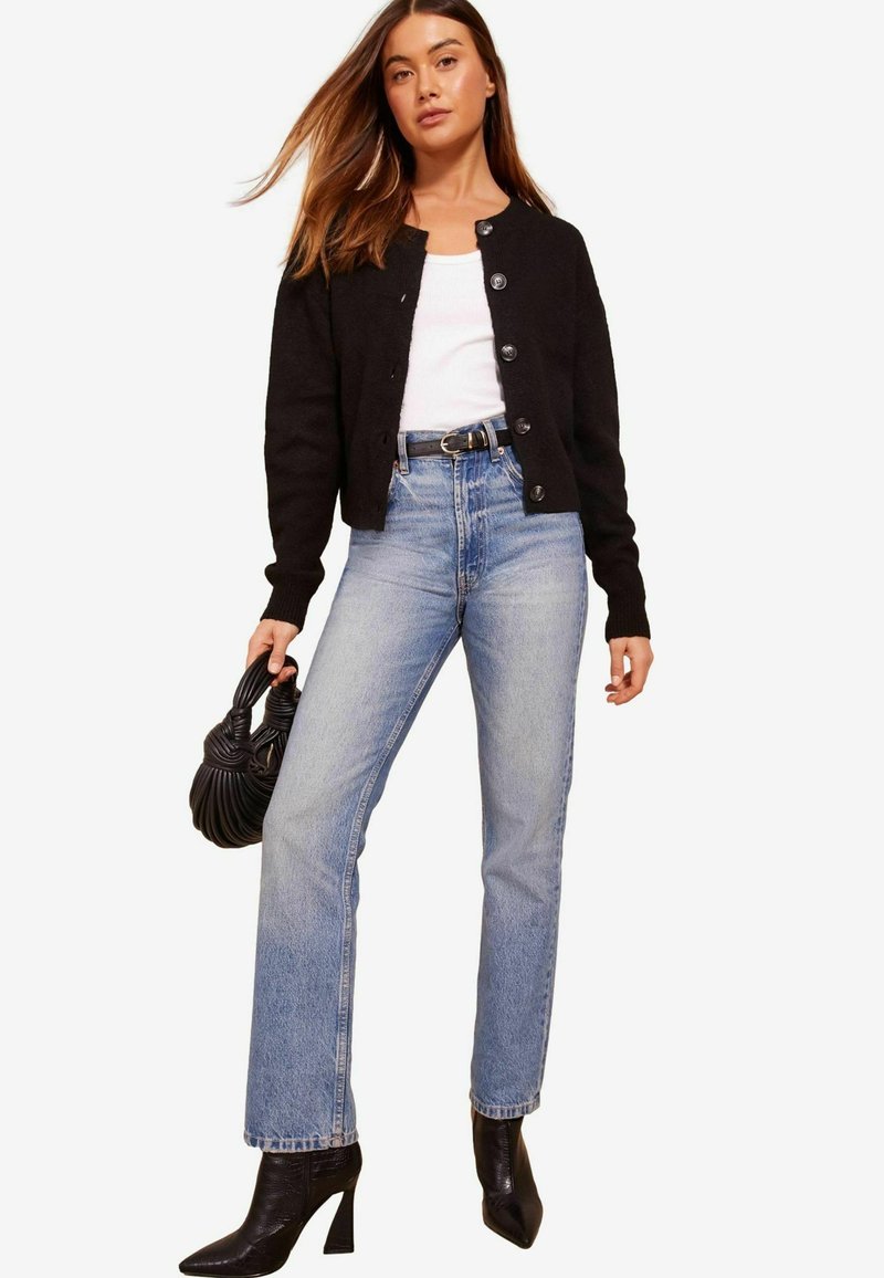 Black knitted cardigan with buttons, white tank top, high-waisted light blue jeans, and black pointed heel boots. Holding a black bag.