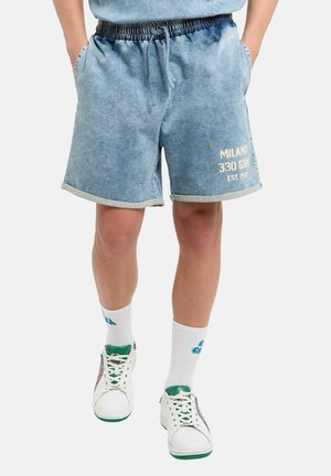 Man draagt blauwe denimshorts met opgerolde zomen, witte sokken en witte sneakers met groene accenten, staand met handen in de zakken.