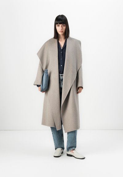 Ženska s ravnimi temnimi lasmi, oblečena v bež oversize plašč, modro srajco, svetlo modre kavbojke, bele loafers čevlje in drži modro torbico.