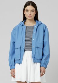 Veste imperméable bleu clair avec capuche, boutons-pression, deux poches avant et poignets élastiques ; portée par-dessus un haut rayé et une jupe blanche.