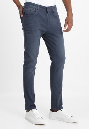 Homme portant un jean bleu foncé ajusté et des baskets blanches, debout avec une main dans la poche devant un fond uni.