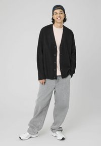 Cardigan noir côtelé avec six boutons, porté sur une chemise rose clair. Associé à un jean gris ample et des baskets blanches. Le chapeau est noir.