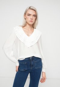 Hvid langærmet bluse med en stor plisseret flæsekrave, parret med blå højtaljede jeans. Glat stof med let glans.