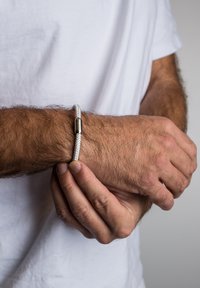 Austi baltas apyrankė su sidabriniu metaliniu akcentu, turinti cilindrinį užsegimą, dėvima ant riešo. Artimas vaizdas, pabrėžiantis tekstūros detales.