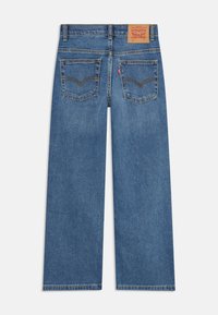 Calças de ganga azul de perna larga com cintura alta, dois bolsos traseiros e um patch de pele da marca na parte posterior da cintura. Textura suave.