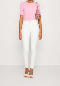 Top ajustado de manga corta rosa, pantalones blancos de cintura alta con cremalleras laterales y sandalias de tiras trenzadas beige. Bolso texturizado al hombro.