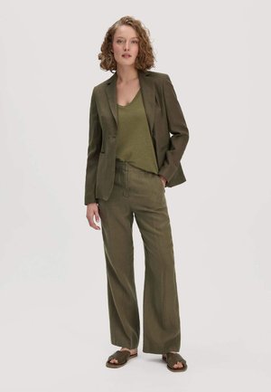 Vrouw met krullend haar draagt een olijfgroene blazer, bijpassende wijde broek, groene top en donkergroene sandalen tegen een effen achtergrond.