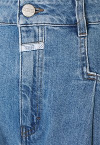 Ljust blå denimjeans har en knappstängning, en etikett med texten "CLOSED" och synlig sömnad längs sömmarna.