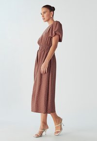 Smeđi midi haljina s točkicama, s napućenim rukavima, elastičnim strukom i lepršavom suknjom; uparena s bijelim sandalama na petu. Glatka tekstura.