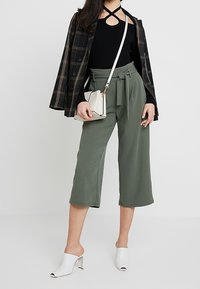 Černý žebrovaný top s křížovými detaily v přední části, kostkovaný blejzr, olivově zelené culottes s mašlí a bílé otevřené muly. Malá kabelka.