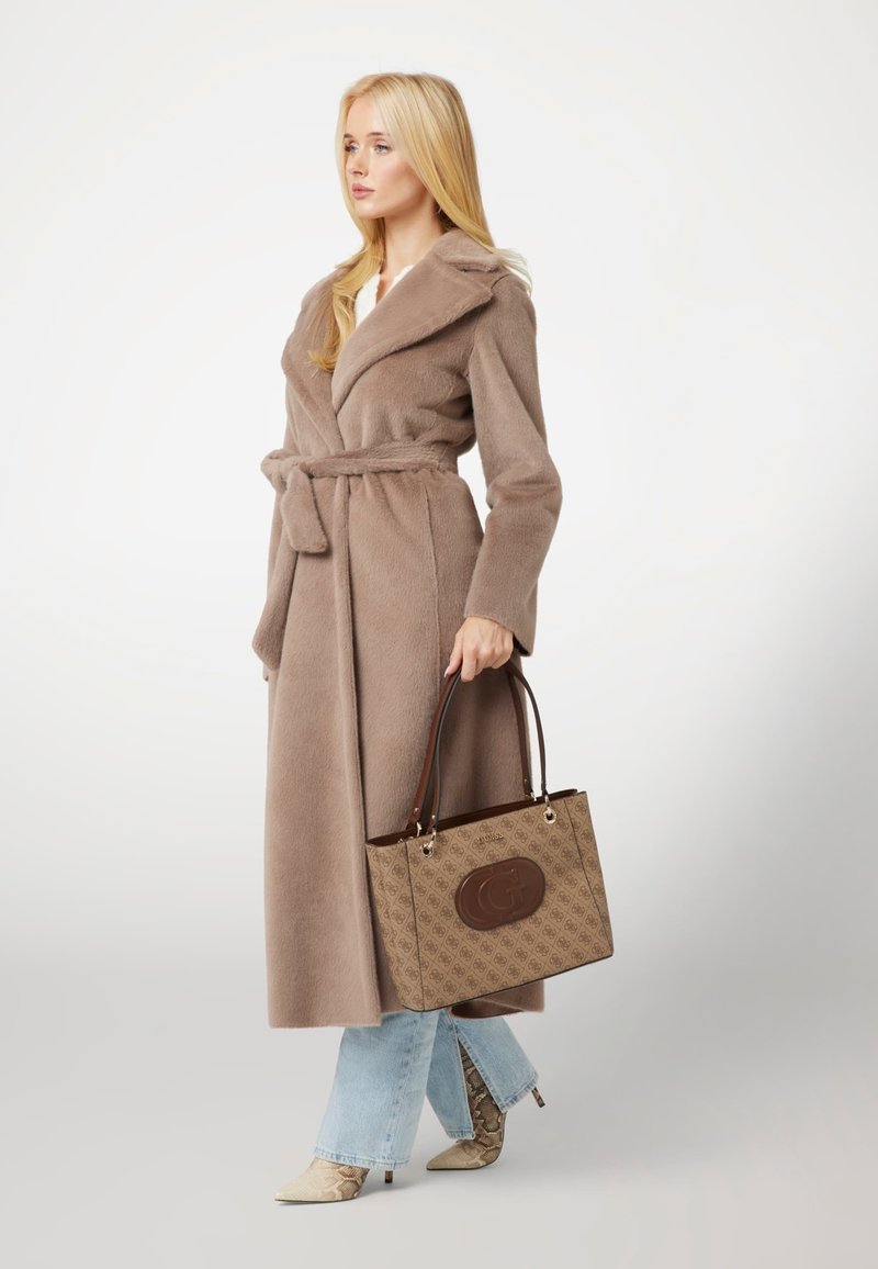 Cappotto in pelliccia sintetica marrone con vita regolabile, jeans svasati azzurri chiari e stivaletti alla caviglia beige in pelle di serpente. Borsa a tracolla marrone con motivo e logo.