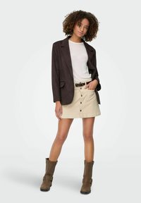 Blazer en daim marron sur un t-shirt blanc, associé à une jupe crème avec des boutons et des poches. Bottines marron à boucles.