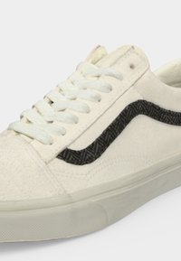 Baskets basses blanches avec des lacets épais et une bande en tissu noire en zigzag sur le côté, avec une semelle en caoutchouc blanche.