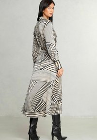 Abito lungo con bottoni, caratterizzato da un motivo geometrici in nero e beige, realizzato in tessuto morbido. Il modello indossa stivali neri fino al ginocchio.