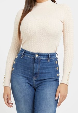 Frau trägt einen cremefarbenen Pullover mit Zopfmuster und langen Ärmeln sowie hoch sitzende blaue Jeans mit goldenen Knöpfen an den Seiten und Ärmeln.