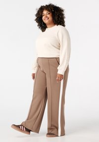 Crème kleurige trui met parelaccenten op de mouwen, gecombineerd met bruin brede broek met witte zijkanten. Bruin en roze gestreepte schoenen.