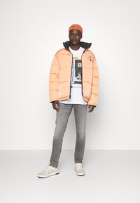 Giacca a vento arancione con rifiniture nere e logo, t-shirt bianca con stampa grafica, jeans grigi e sneaker beige con accenti arancioni.