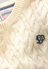 Cremefarbener, kabelgefütterter Pullover mit Rundhals, detaillierter Textur und kleinem gestickten Logo auf der Vorderseite.