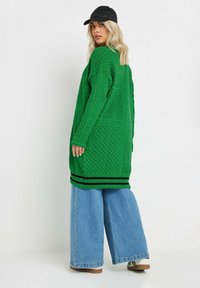 Grön stickad cardigan med mönster, långa ärmar och svarta randiga detaljer vid fållen. Tillsammans med vida blå denimjeans.