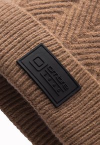 Gorro de punto marrón con un patrón texturizado en zigzag, que presenta un parche rectangular negro embosado con "ombre 1985."