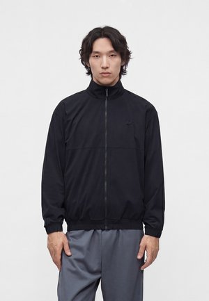 Hombre con cabello oscuro de longitud media, vestido con una chaqueta negra con cremallera y pantalones grises holgados, de pie frente a un fondo blanco liso.