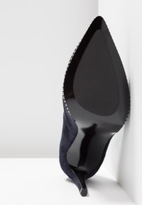 Talons aiguilles en suède bleu marine avec un bout pointu, présentant un dessous noir élégant et des accents argentés le long du bord.