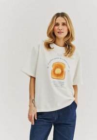 Bílý bavlněný tričko s krátkým rukávem, zdobené potiskem toastu a textem. Design zahrnuje teplé barvy a volnější střih.