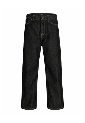 Adolescent aux cheveux bouclés portant une chemise marron, une surchemise à carreaux rouges, un jean ample foncé et des bottes couleur sable, debout devant un fond uni.