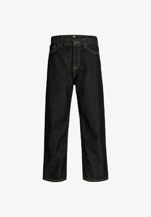 Adolescent aux cheveux bouclés portant une chemise marron, une surchemise à carreaux rouges, un jean ample foncé et des bottes couleur sable, debout devant un fond uni.
