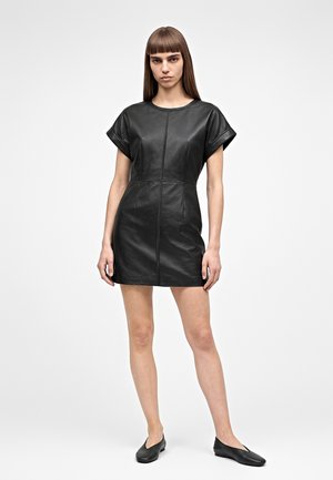 Femme debout portant une mini-robe en cuir noir à manches courtes et des chaussures plates noires sur un fond gris clair.