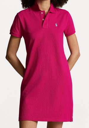 Kobieta ubrana w różową sukienkę polo z krótkim rękawem, z małym, jasnoniebieskim haftowanym logo na piersi.