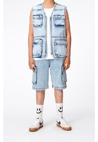 Džínová vesta s několika kapsami, přes bílé tričko, spárovaná s džínovými šortkami po kolena. Obsahuje ponožky se smajlíkem a tenisky.