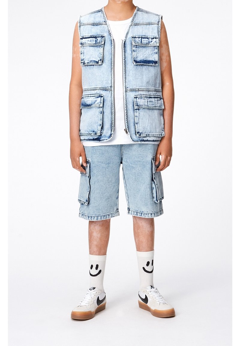 Džínová vesta s několika kapsami, přes bílé tričko, spárovaná s džínovými šortkami po kolena. Obsahuje ponožky se smajlíkem a tenisky.