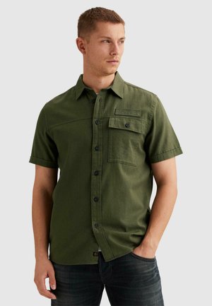 Muž v olivovo zelenej košeli s krátkymi rukávmi a náprsným vreckom, stojaci s jednou rukou v vrecku riflí, pozerajúci sa do strany.