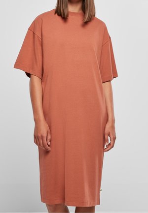 Kobieta ubrana w luźną, krótko rękawową, sięgającą kolan sukienkę w kolorze terakoty, stojąca na tle jednolitego jasnoszarego tła.