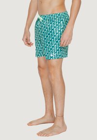 Costumi da bagno verdi con un motivo ondulato bianco, realizzati in materiale leggero. Presentano una vita elasticizzata con cordoncino.