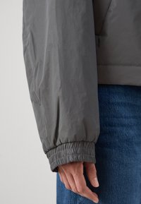 Grå, lett jakke med elastiske mansjetter, med en glatt tekstur og en litt glanset finish, kombinert med lyseblå jeans.