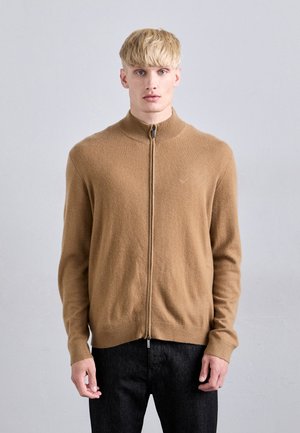 Suéter marrón con cremallera, hecho de un tejido suave, que presenta un cuello alto, puños acanalados y una textura lisa. Lleva puesto con unos jeans oscuros.