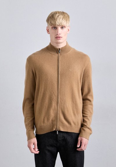 Maglione marrone zip-up realizzato in tessuto morbido, con collo alto, polsini a coste e una superficie liscia. Indossato sopra jeans scuri.