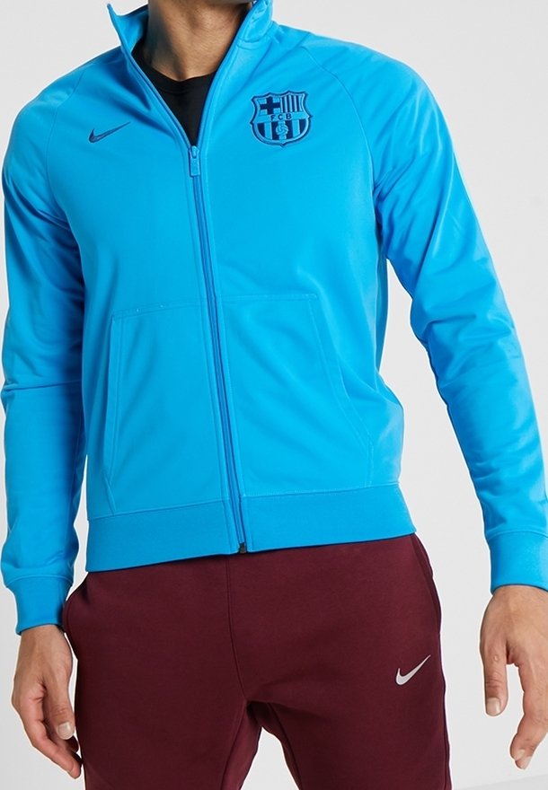 Hellblaues Zip-Jackett mit Barcelona-Logo, hergestellt aus glattem Material, mit zwei Fronttaschen und eng anliegenden Bündchen.