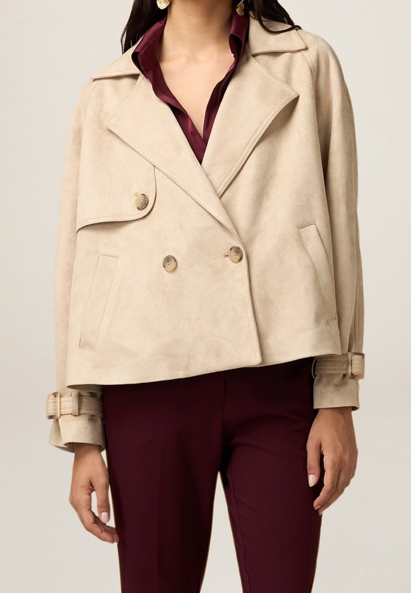 Veste courte en daim beige à double boutonnage avec de gros boutons, col cranté et poignets de manches ceinturés. Associée à un pantalon bordeaux foncé.