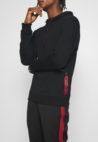 Hoodie noir avec une fermeture éclair à l'avant et un accent de logo rouge sur le côté. Fabriqué en tissu lisse avec une coupe décontractée et une capuche réglable à cordon.