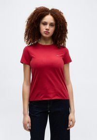 Νεαρή γυναίκα με σγουρά μαλλιά που φοράει κόκκινο κοντομάνικο μπλουζάκι και σκούρο τζιν, στέκεται μπροστά σε απλό φωτεινό φόντο.